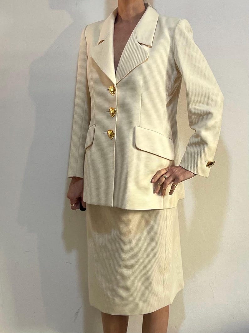 Puede incluir: Traje de falda color crema. La chaqueta tiene solapas con muescas, tres botones dorados en forma de coraz&oacute;n y un bolsillo. La falda llega a la rodilla. El traje tiene una tela acanalada texturizada.