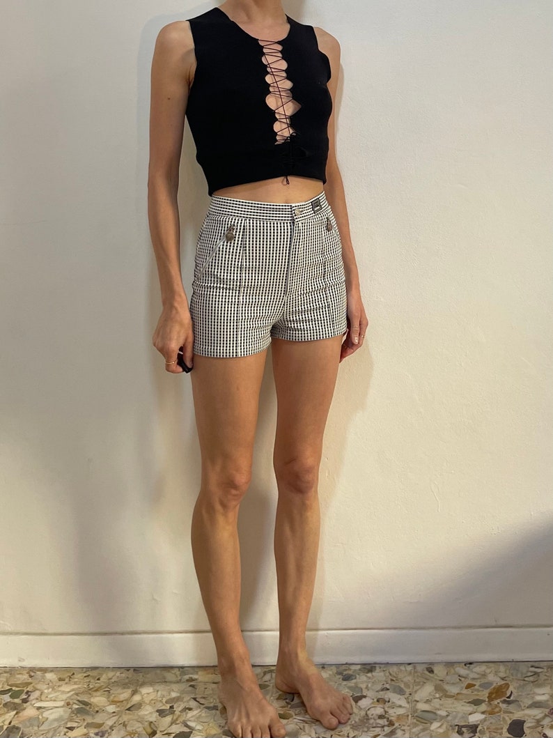 May include: Black sleeveless top with a lace-up front and high-waisted, black and white checkered shorts. The shorts have two decorative buttons on the front. The outfit is a stylish ensemble.