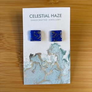 Op de afbeelding: Een paar vierkante, blauwe oorstekers met gouden adering, gepresenteerd op een kaart met de tekst "Celestial Haze Handcrafted Jewellery". De kaart heeft een aquarelontwerp in blauw, groen en goud.