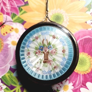 May include: A round pendant featuring a central basket of flowers motif in pink, blue, and green enamel on a white guilloché background. The design is framed by a light blue border and a dark outer rim, suspended from a thin silver-toned chain.