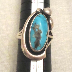 May include: A silver ring featuring a large, oval turquoise stone with dark brown inclusions. The turquoise is set in a silver bezel with a decorative silver design. The ring is on a black ring sizer.
