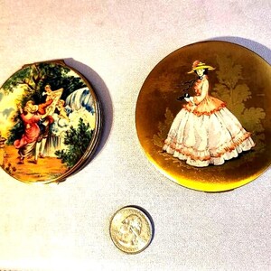 May include: Two antique compacts with painted scenes. One depicts a romantic outdoor scene with figures near a waterfall. The other features a woman in a long dress and hat. A quarter is included for size reference.