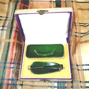 May include: A rectangular presentation box with a white interior and purple trim containing two dark green items. One item is a rectangular brush with a curved handle, and the other is a shoehorn with a metal hook attached to a green handle.