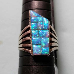 May include: A silver ring featuring a rectangular inlay of iridescent blue opal tiles. The ring has a multi-band shank design that splits into four separate silver wires on each side, supporting the central stone setting with a flat top edge.