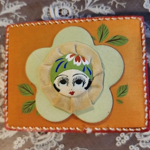 May include: An orange fabric box with a light green flower design and a doll face in the center. The box has a red and white rope trim and green leaves painted on the orange background.