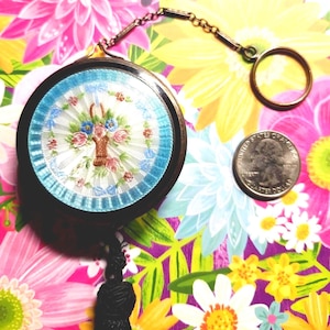 May include: A round compact mirror with a black frame and a central floral embroidery featuring a basket of pink and blue flowers on a white background, surrounded by a blue border. It includes a metal chain, a ring, and a black tassel.