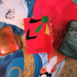 May include: A red diary with a black and green design, a green and black marbled clutch, and a red and orange marbled clutch. The diary has the word "DIARY" printed on the front.