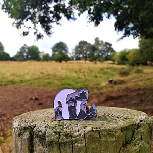 May include: A purple and black enamel pin depicting Stonehenge, a prehistoric monument in England. The pin is set against a blurred background of a field and trees.