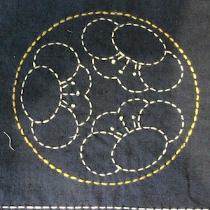 May include: A dark blue fabric square with a white and yellow stitched design. The design is a circle with a series of overlapping circles inside.