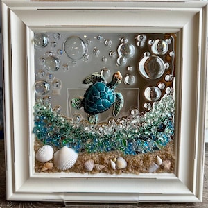May include: A framed artwork featuring a teal sea turtle swimming amidst bubbles and sea glass. The artwork includes seashells and sand, creating a beach scene. The frame is white.