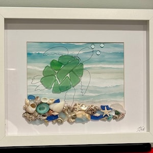 May include: A framed artwork featuring a sea turtle made of green sea glass. The turtle swims in front of a watercolor ocean scene. The base of the artwork is adorned with seashells, starfish, and blue glass pieces. The frame is white.