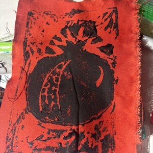 May include: A square piece of red fabric with a black pomegranate print. The pomegranate is centered on the fabric, with a detailed design. The edges of the fabric are frayed.