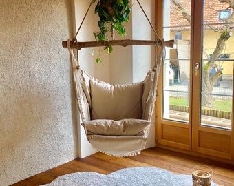 Könnte beinhalten: Ein weißer Hängesessel mit einem beigefarbenen Kissen und einem weißen, flauschigen, wolkenförmigen Teppich. Eine Topfpflanze hängt von der Decke über dem Sessel.