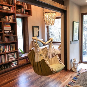 Puede incluir: Una silla hamaca verde cuelga del techo en una habitación con suelo de madera y una estantería. La silla hamaca está hecha de cuerda trenzada y tiene un asiento de tela verde. La habitación está decorada en un estilo rústico con una viga de madera y una lámpara de araña.