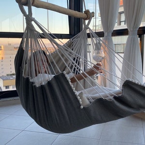 Puede incluir: Una persona relajándose en una hamaca gris y blanca con una copa de vino. La hamaca está colgada de una barra de madera y está suspendida frente a una gran ventana con vista a la ciudad.