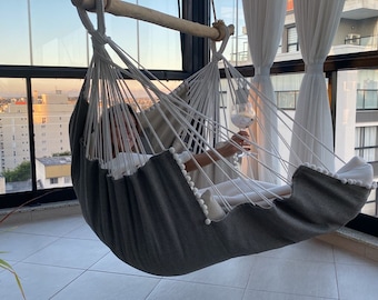 May include: A person relaxing in a gray and white hammock with a glass of wine. The hammock is hanging from a wooden bar and is suspended in front of a large window with a city view.