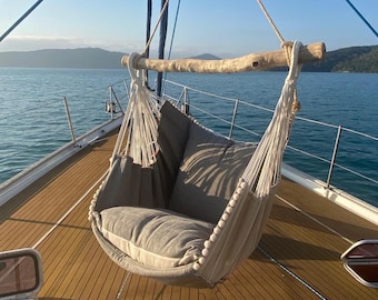 May include: A white hammock with beige cushions hangs from a wooden beam on a boat deck. The boat is docked in a harbor with blue water and green hills in the background.