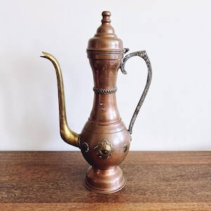 May include: A copper-colored vintage teapot with a long, curved spout and ornate handle. The teapot features decorative brass accents and a lid with a small knob. The design is reminiscent of Middle Eastern or antique styles.