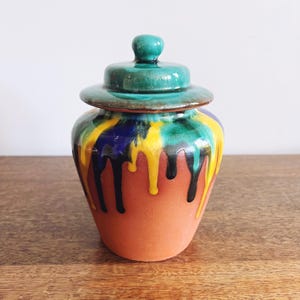 May include: A ceramic jar with a lid, featuring a terracotta base and a vibrant, dripped glaze design. The glaze includes yellow, blue, green, and black colors. The lid is a matching green color.