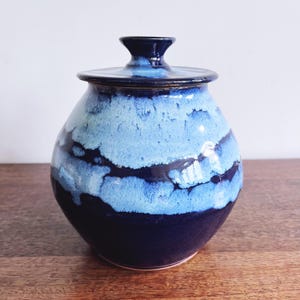 May include: A ceramic jar with a lid, featuring a gradient of blues. The jar's base is a deep navy, transitioning to lighter shades of blue with a textured, cloud-like pattern. The lid and finial are a darker navy.