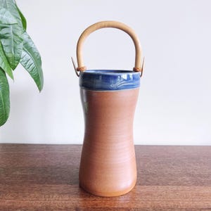 May include: A handcrafted ceramic vase with a brown body and a blue rim. It features a curved wooden handle attached with leather straps. The vase has a unique, organic shape and is suitable for displaying flowers or as a decorative piece.