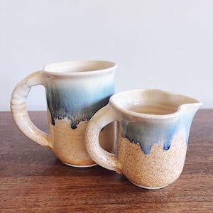 May include: A ceramic mug and a matching creamer pitcher featuring a speckled tan base and a blue drip glaze finish. Both pieces have thick, textured handles and a smooth interior. The pitcher has a distinct pouring spout on the rim.