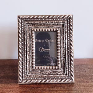 May include: Ornate, rectangular photo frame with a cream and gold finish. The frame features a rope-like border and detailed molding. The text "Vintage Editions" and "Rama PHOTO FRAME" are visible. The frame holds a 4" x 6" photo.