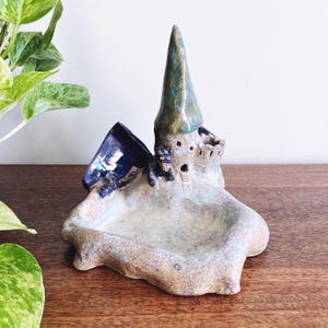 May include: A ceramic trinket dish featuring a sculpted castle with a tall, pointed blue-green roof, a small dark blue bowl attachment, and a textured, sandy-colored base. The structure includes miniature windows and a small arched doorway.