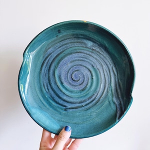 May include: A handmade ceramic plate with a teal and blue spiral design. The plate has a glossy finish and a unique shape with a small handle on the side. The plate is perfect for serving appetizers or as a decorative piece.