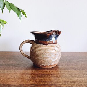 May include: A brown and white ceramic pitcher with a blue glaze. The pitcher has a rounded body and a handle.