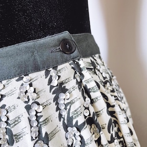May include: Close-up of a skirt with a patterned design. The skirt features a gray waistband with a button closure. The fabric has a white background with a dark gray abstract pattern and is embellished with sequins.