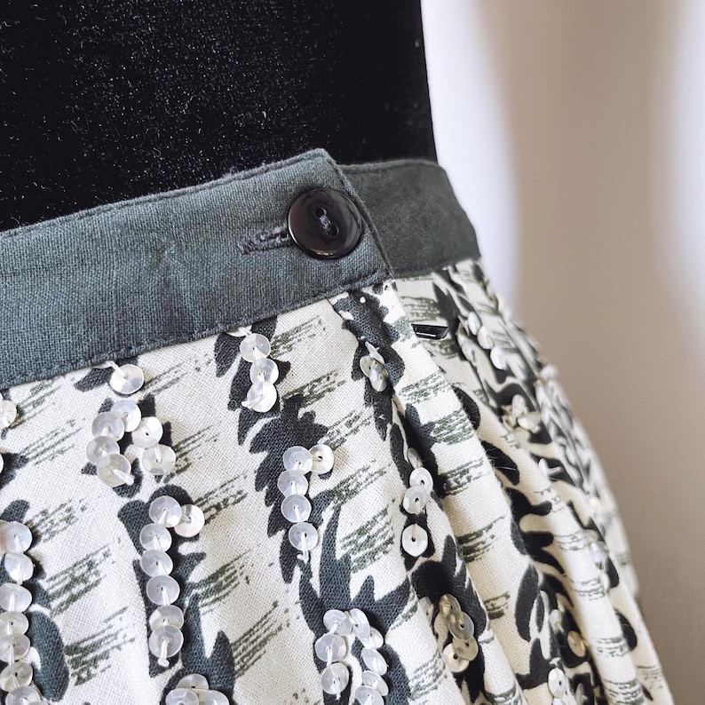 May include: Close-up of a skirt with a patterned design. The skirt features a gray waistband with a button closure. The fabric has a white background with a dark gray abstract pattern and is embellished with sequins.