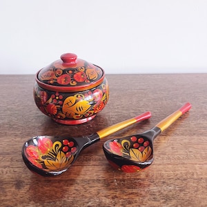 May include: A hand-painted wooden sugar bowl with a lid and two matching spoons. The bowl and spoons are black with red, yellow, and orange floral designs. The spoons have gold handles.