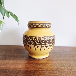 May include: A yellow ceramic vase with a brown geometric pattern. The vase has a wide mouth and a rounded body.