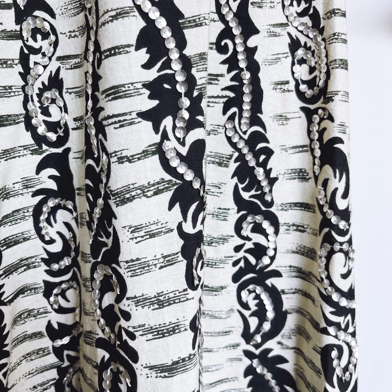 May include: Close-up of a fabric with a black and white pattern. The fabric features vertical stripes with a black floral design and silver sequins. The background has a white base with horizontal black brushstrokes.