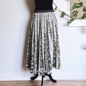 May include: A long, pleated skirt with a black and white pattern. The skirt features vertical stripes with a repeating floral design. The waistband is black, and the skirt is displayed on a black mannequin stand. The skirt is floor length.