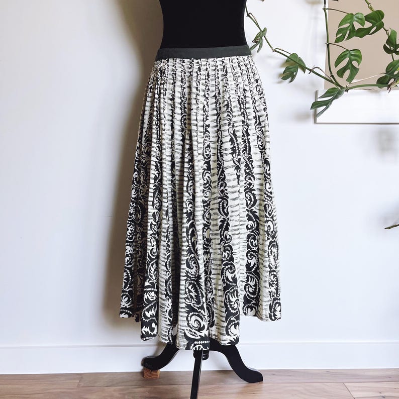 May include: A long, pleated skirt with a black and white pattern. The skirt features vertical stripes with a repeating floral design. The waistband is black, and the skirt is displayed on a black mannequin stand. The skirt is floor length.