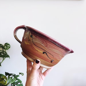 May include: A large, handmade ceramic mixing bowl with a brown, red, and white glaze. The bowl has a unique marbled design and a single handle.