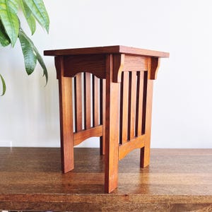 May include: A square, wooden side table with a warm, reddish-brown finish. The table features a slatted design on the sides and a solid top. The legs are sturdy and the overall style is reminiscent of Arts and Crafts furniture.