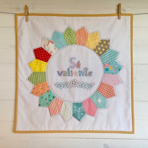 Puede incluir: Un cuadrado de tela blanco con borde amarillo, que presenta un diseño de flor de patchwork colorido con la frase en español "Sé valiente" bordada en el centro.