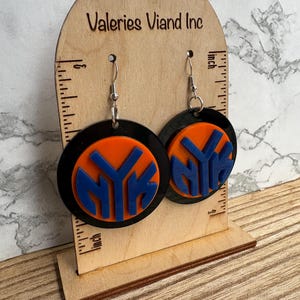 May include: A pair of round earrings featuring the letters "NYK" in blue, set against an orange background, and framed by a black circle. The earrings are displayed on a wooden stand with the text "Valeries Viand Inc".