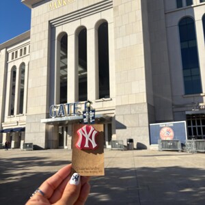 May include: A red and white New York Yankees logo pin attached to a rectangular brown cardboard display card. The card features the text ValerieVandInc and Made in The Bronx, NY. The background shows the exterior facade of Yankee Stadium.