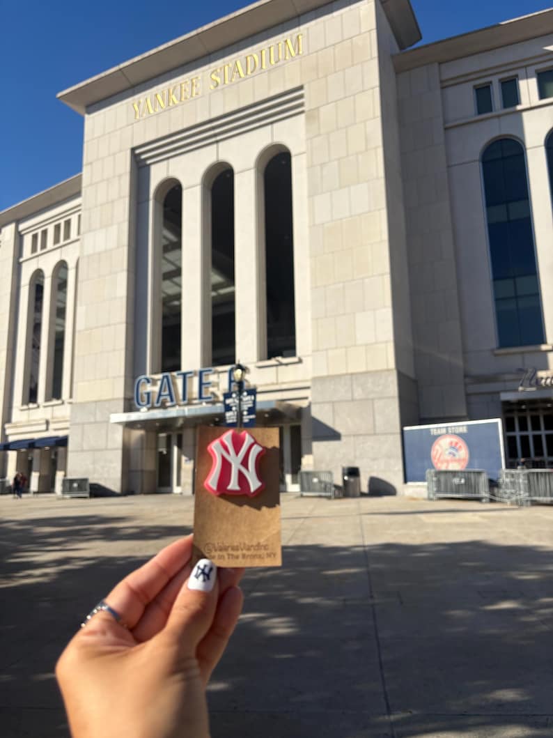 May include: A red and white New York Yankees logo pin attached to a rectangular brown cardboard display card. The card features the text ValerieVandInc and Made in The Bronx, NY. The background shows the exterior facade of Yankee Stadium.