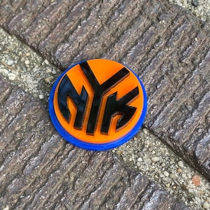 May include: A round, orange and blue pin with a black "NY" logo. The pin is on a textured surface.