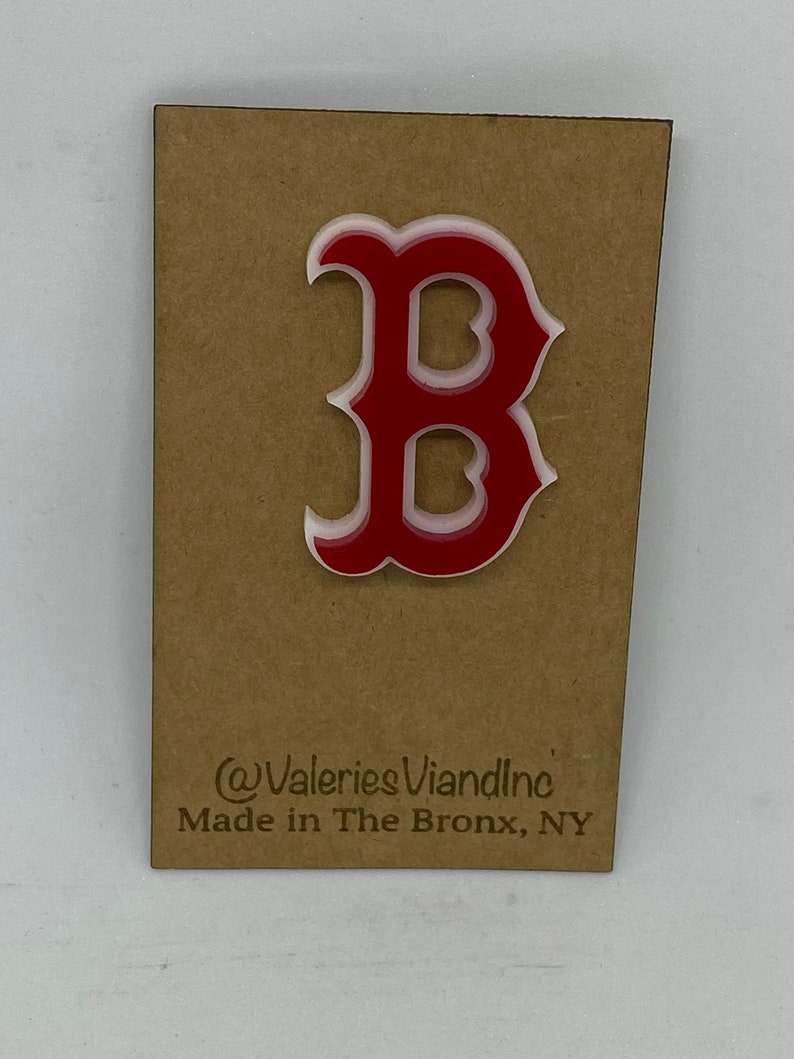 May include: A red and white acrylic letter B pin attached to a rectangular brown cardboard backing. The backing features the text @ValeriesViandInc and Made in The Bronx, NY printed in black ink at the bottom of the display card.
