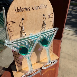 May include: Clear acrylic martini earrings with green olive accents. The earrings are displayed on a wooden stand with the text "Valeries Viand Inc". The earrings are approximately 7.6 cm long.