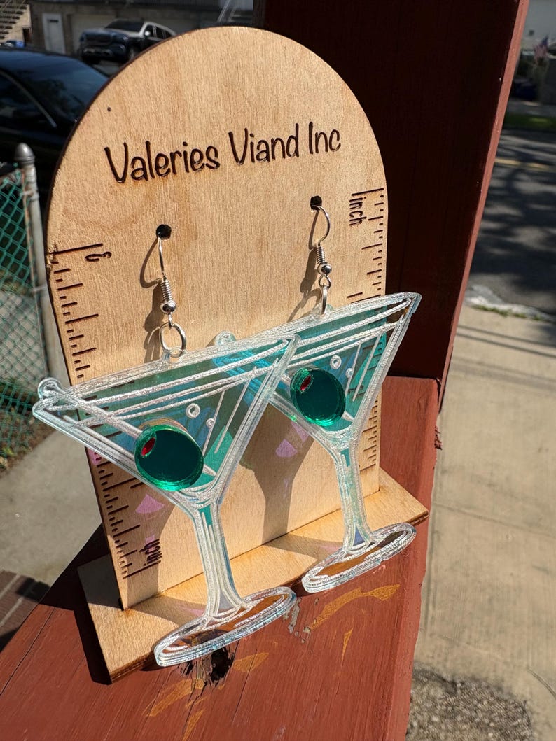 May include: Clear acrylic martini earrings with green olive accents. The earrings are displayed on a wooden stand with the text "Valeries Viand Inc". The earrings are approximately 7.6 cm long.