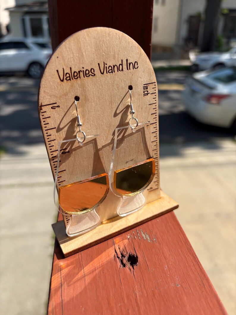 May include: Clear acrylic earrings shaped like whiskey glasses, with a gold-coloured liquid effect. The earrings hang from silver-coloured hooks. The earrings are displayed on a wooden stand with the text "Valeries Viand Inc".