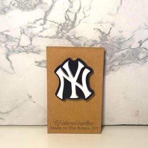 May include: A New York Yankees logo pin featuring a white interlocking NY design on a dark blue background. The pin is attached to a rectangular brown card with the text @ValeriesViandInc and Made in The Bronx, NY printed at the bottom.