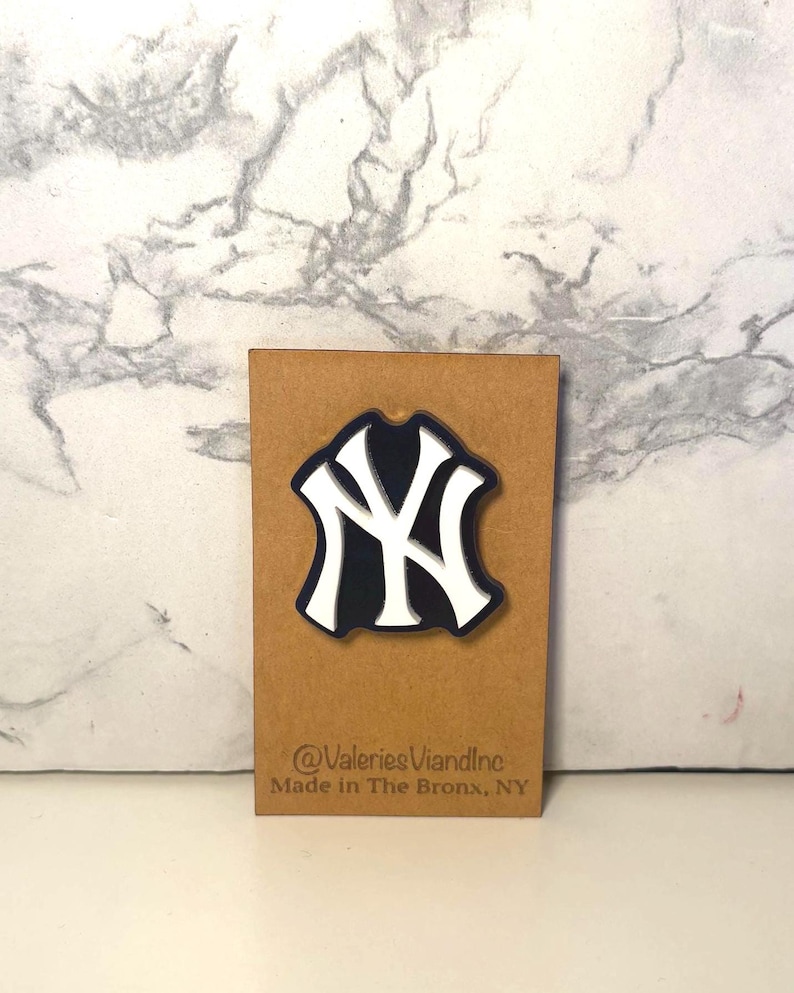 May include: A New York Yankees logo pin featuring a white interlocking NY design on a dark blue background. The pin is attached to a rectangular brown card with the text @ValeriesViandInc and Made in The Bronx, NY printed at the bottom.